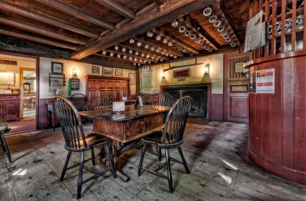 Historic tavern interior with a wooden table, chairs, exposed beams, fireplace, and mugs hanging from the ceiling.
