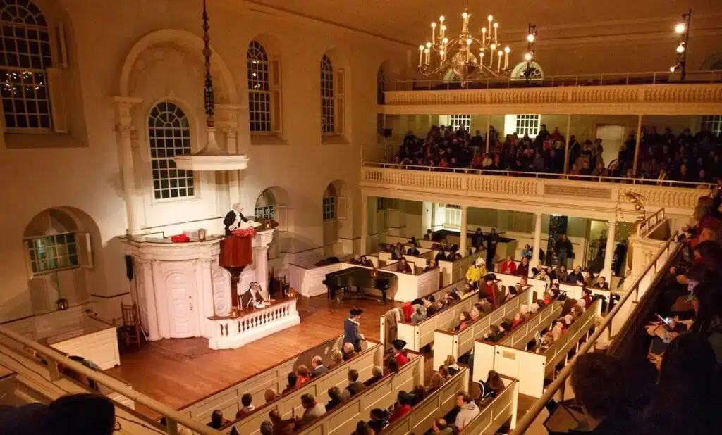 Audience in a church-style hall watches a speaker at a pulpit, with balconies and chandeliers overhead.