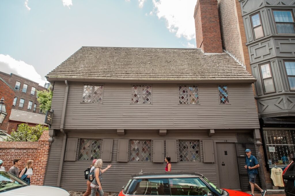 People walk past a historic two-story wooden house with diamond-pane windows on a sunny day.