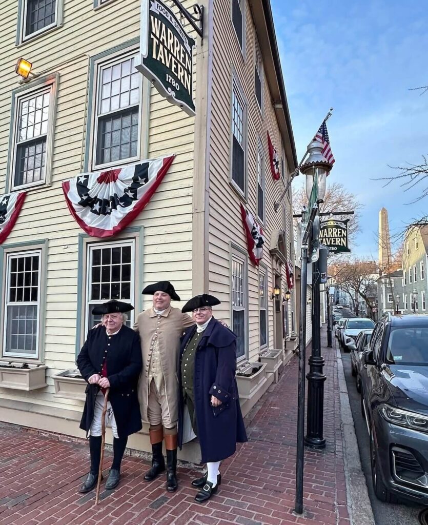 Three people in colonial attire stand outside Warren Tavern decorated with patriotic bunting, on a brick sidewalk.