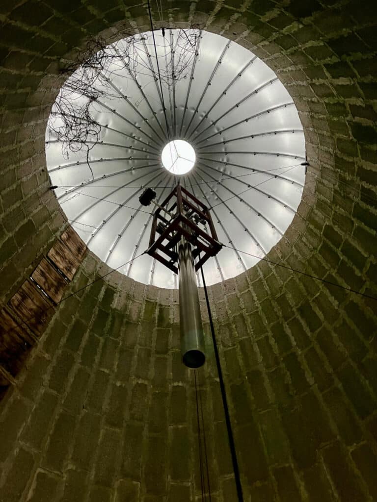 Looking up inside a round, brick-walled silo with a metal roof and central hanging bell made by Paul Matisse for Otto Piene.