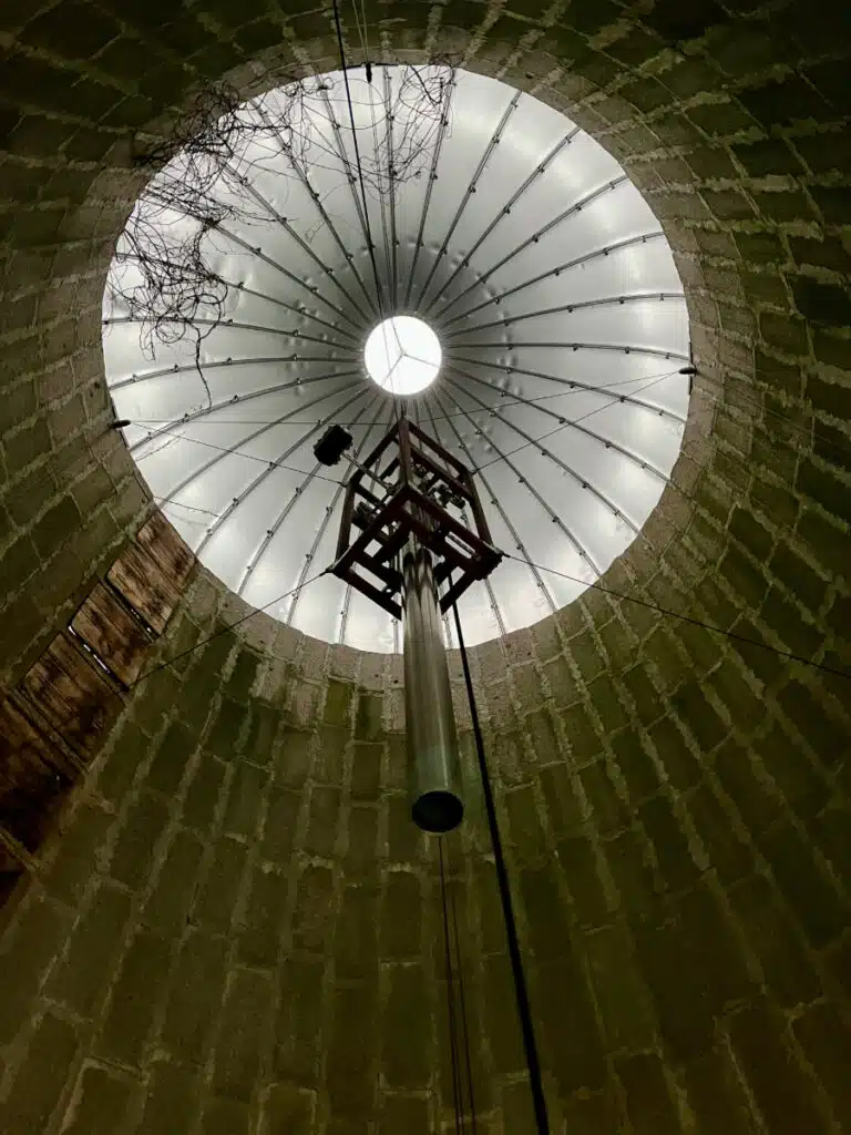 Looking up inside a round, brick-walled silo with a metal roof and central hanging bell made by Paul Matisse for Otto Piene.