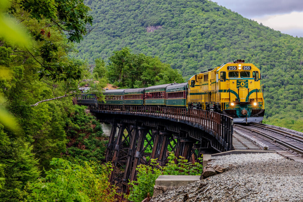 Best New England Summer Attractions for Families. A yellow and green train crosses a curved bridge surrounded by lush green mountains and trees.