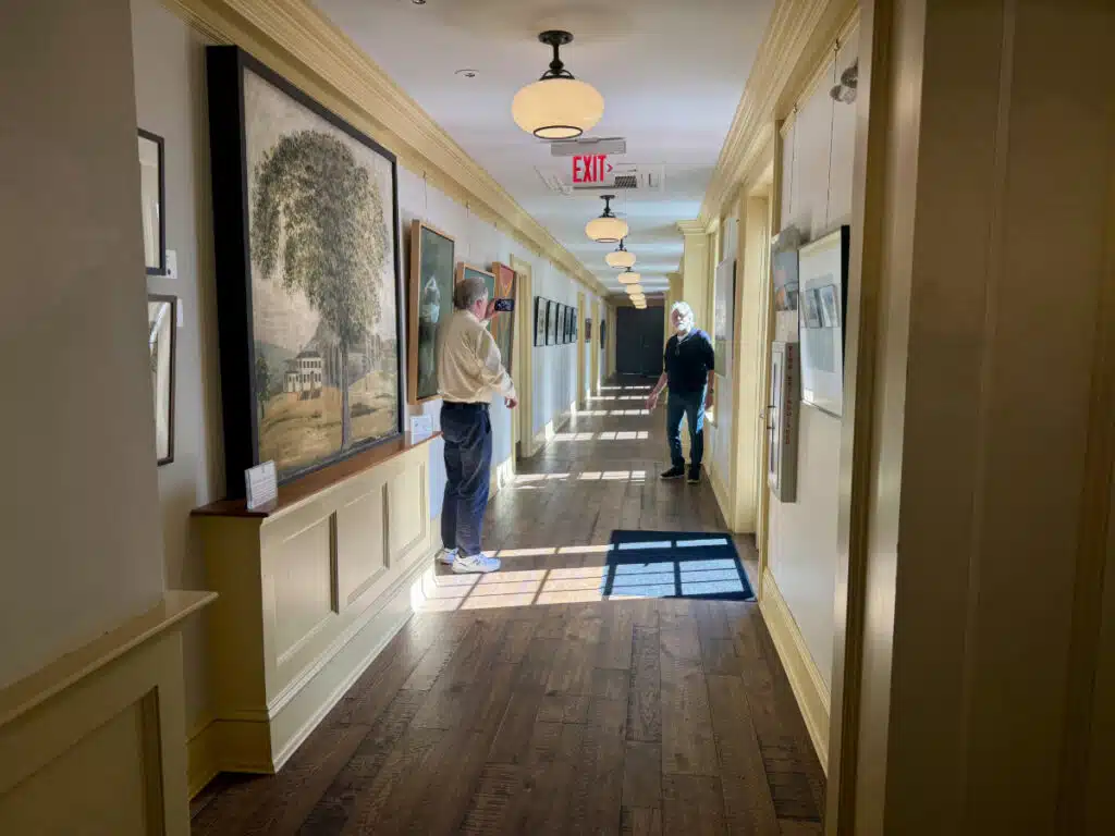NOA Gallery at The Groton Inn. Two men stand in a sunlit hallway with wooden floors and framed artwork on the walls.