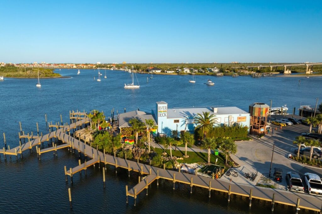River Deck Tiki Bar and Restaurant in New Smyrna Beach, with docks, boats, and palm trees along a wide river under a clear blue sky.