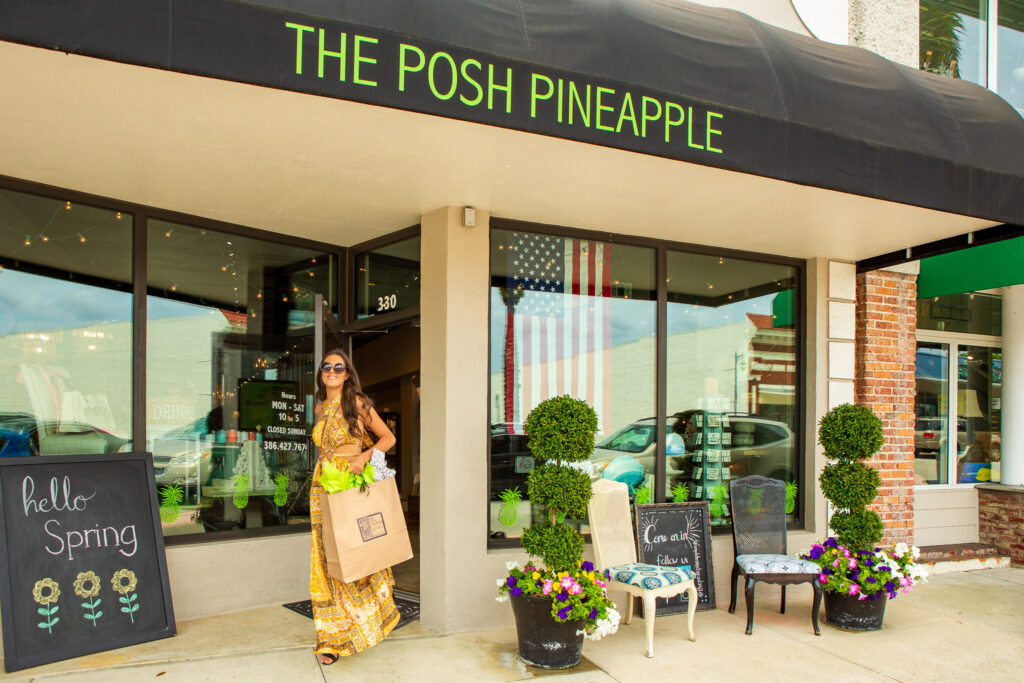 Woman in a yellow dress leaves The Posh Pineapple boutique holding a shopping bag and flowers on a sunny day.