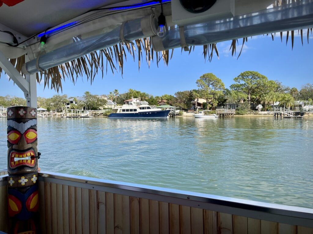 View from a Toon Tiki boat in New Smyrna Beach with a tiki mask, looking out at a river and docked boats under a clear blue sky.