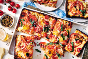 Weeknight Detroit-Style Tomato Pie. Rectangular tomato and basil pizza cut into squares on a tray, with fresh tomatoes and spices on the side.
