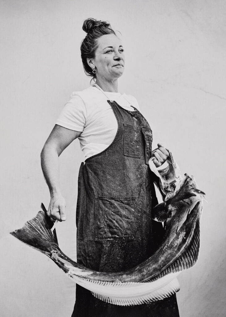 Woman in apron holding a large fish, standing against a plain background, black and white photo.