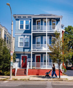 A person pushes a stroller past a blue three-story house with red doors on a sunny day.