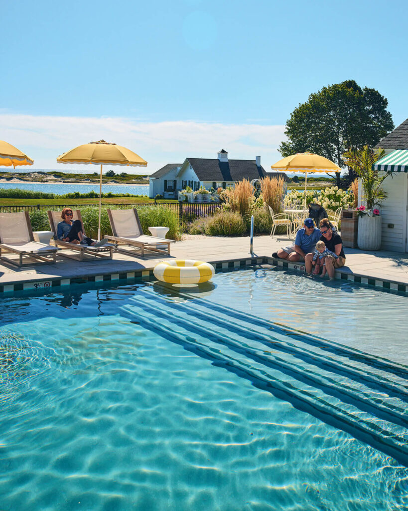 People relax by a sunlit outdoor pool with umbrellas, loungers, and a scenic view of a house and water in the background.
