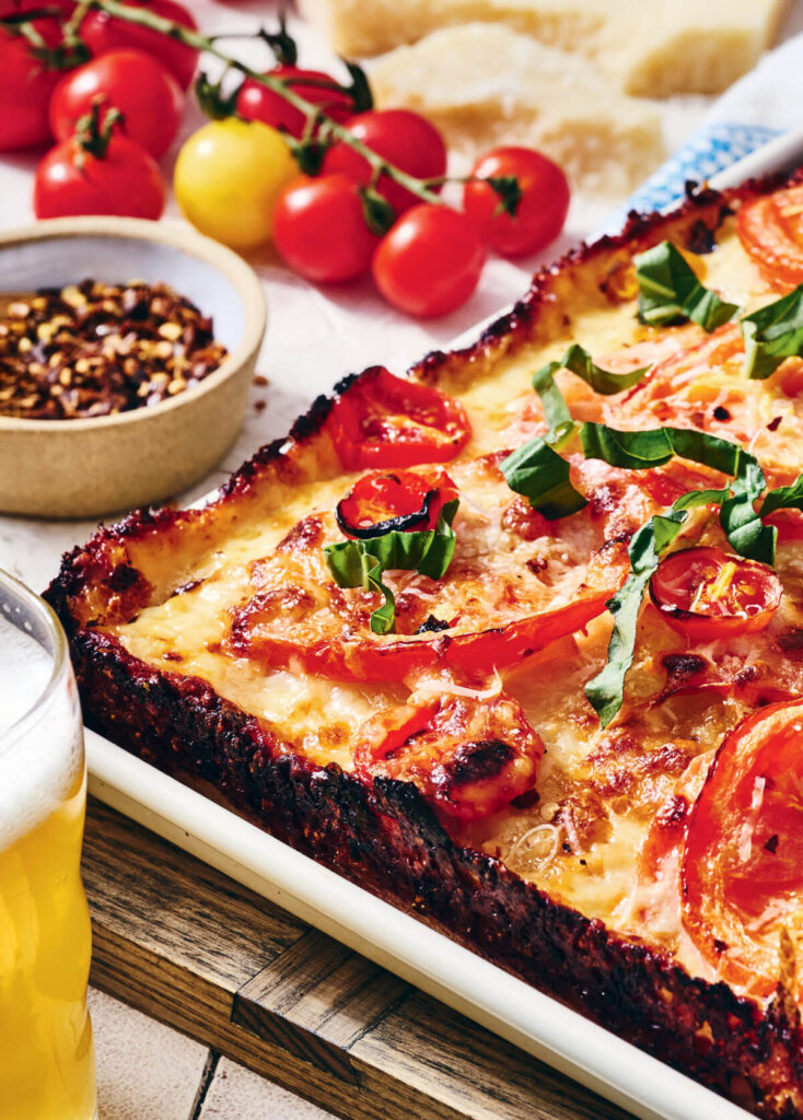 Rectangular pizza with tomatoes and basil, next to a glass of beer, cherry tomatoes, and a bowl of chili flakes.