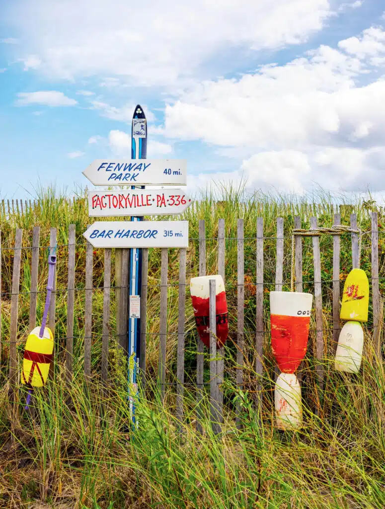 A wooden signpost with distance markers stands by a fence with buoys and tall grass under a partly cloudy sky.
