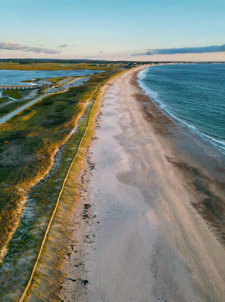 Aerial view of a sandy beach with grassy dunes and blue ocean under a clear sky at sunset.