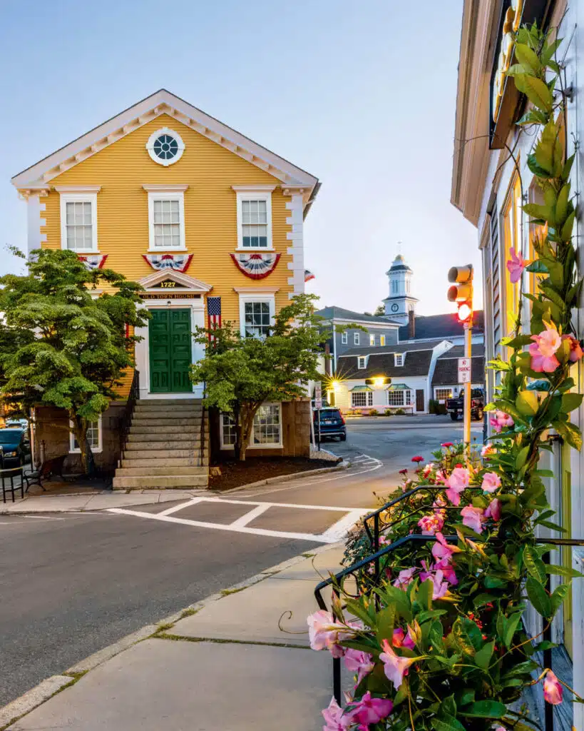 Yellow historic building with green door, red-white bunting, flowers, and a town clock tower in the background.