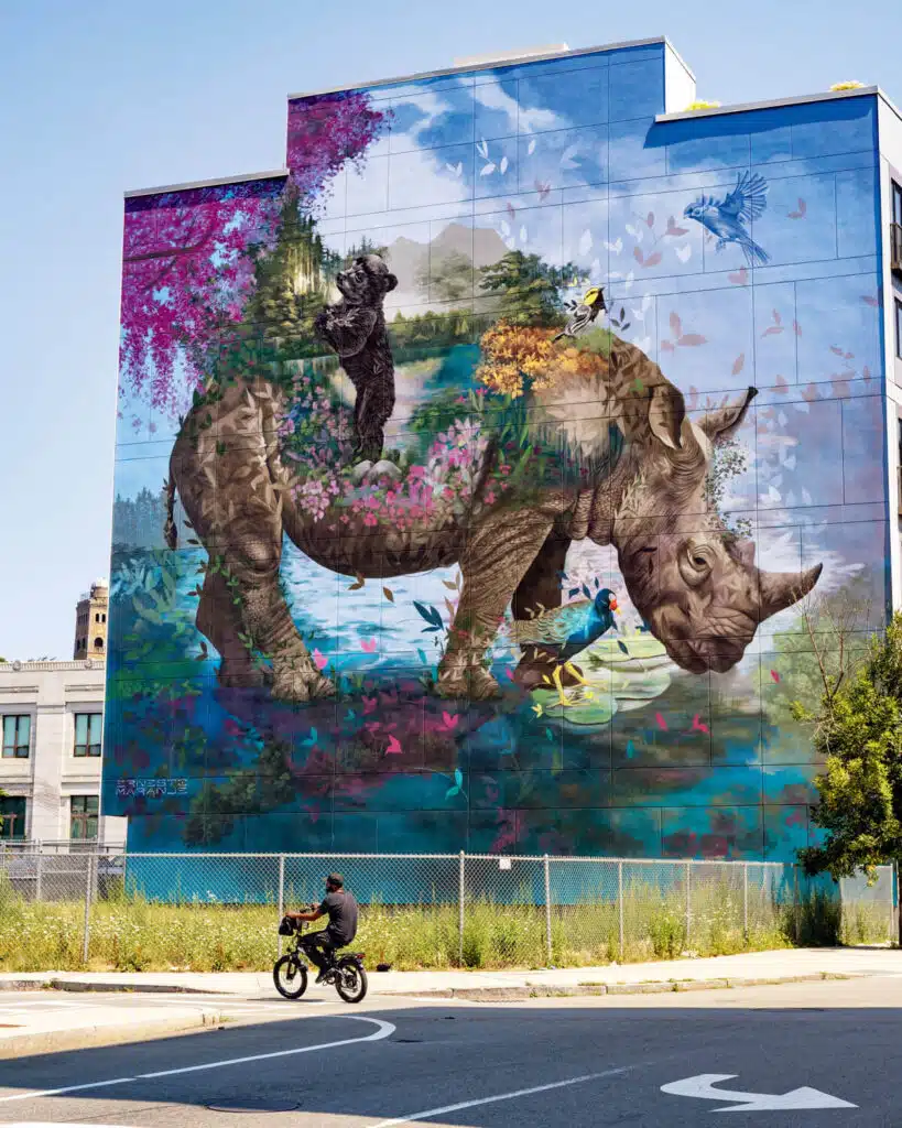 A vibrant mural of a rhino with animals and flowers on its back covers a building wall; a person rides a bike below.