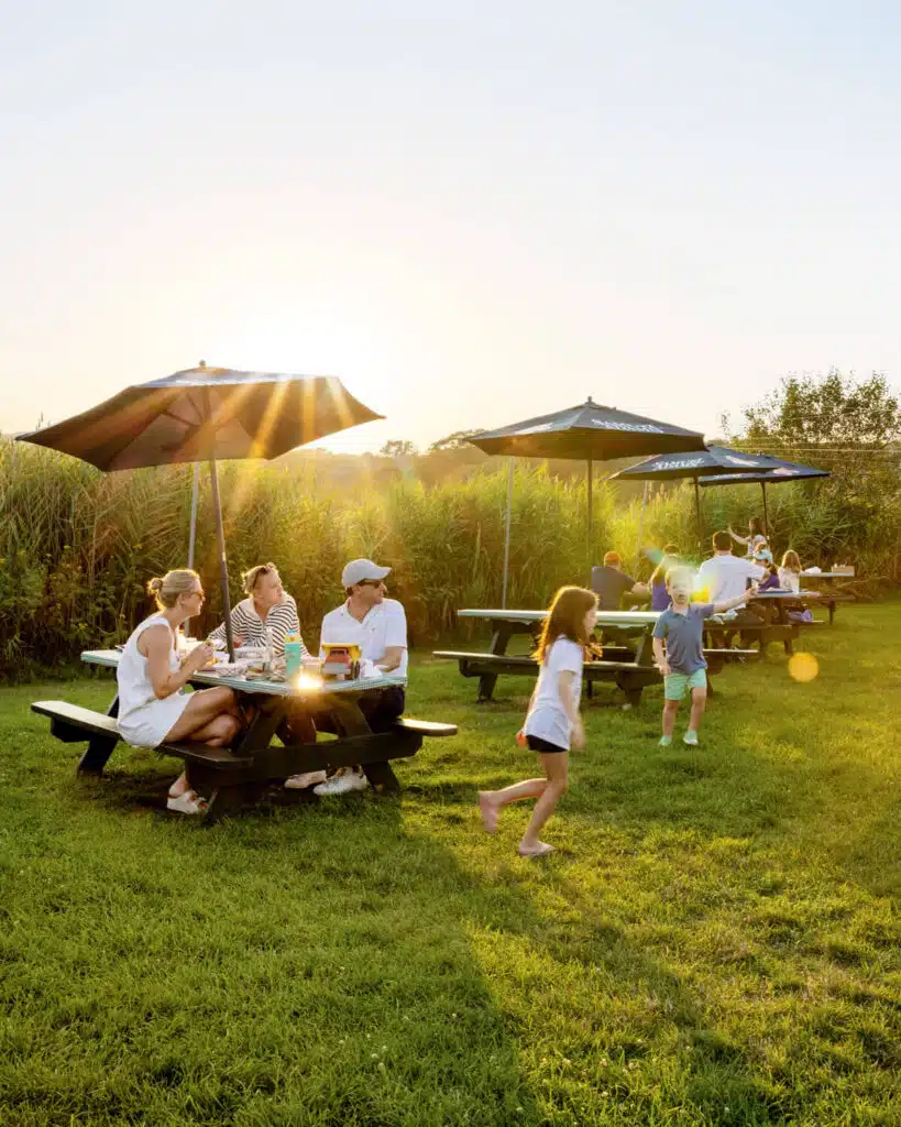 People sitting at picnic tables under umbrellas on a sunny day, with children playing on the grass nearby.
