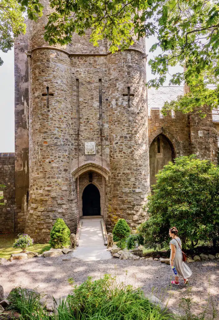 A woman walks toward a large stone castle entrance surrounded by greenery on a sunny day.