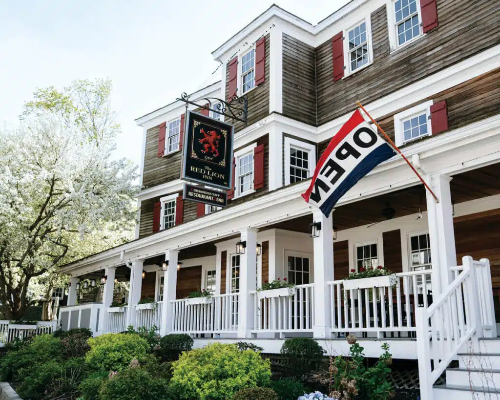 Historic inn with a porch, “OPEN” flag, and “Red Lion Inn” sign, surrounded by greenery and flowers.