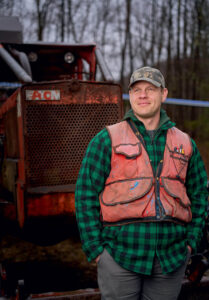 A man in a plaid shirt and orange vest stands in front of a rusty tractor in a wooded area.