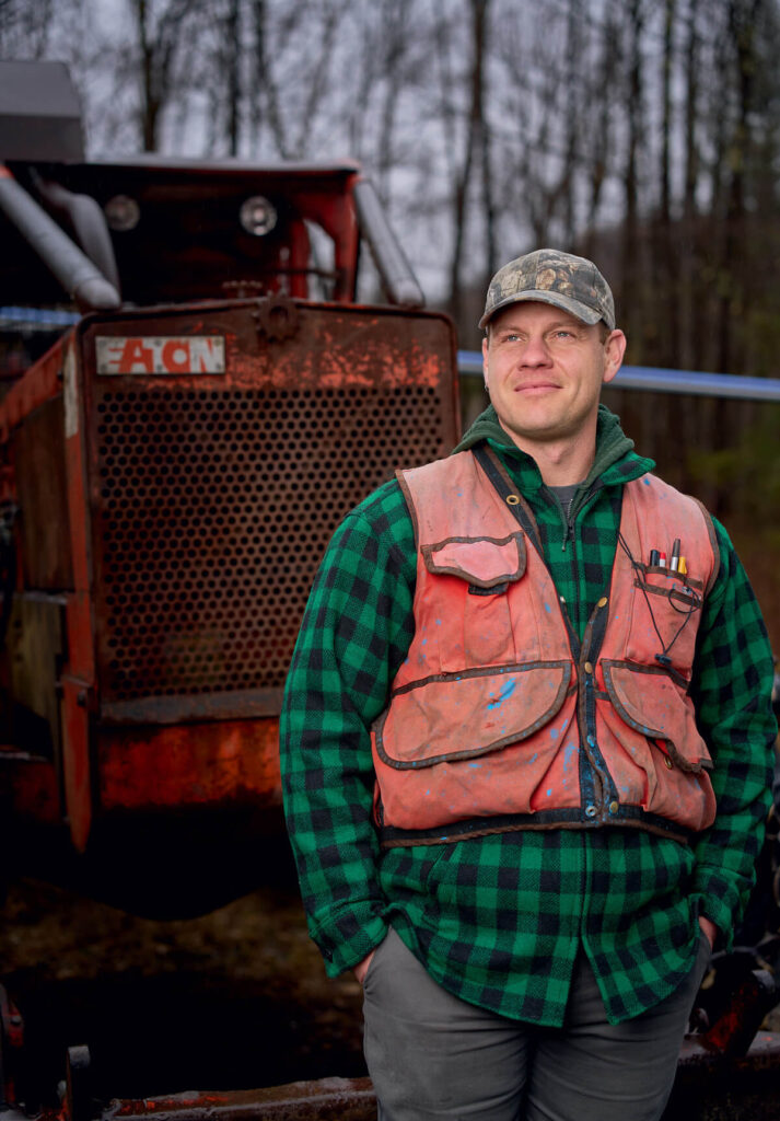 A man in a plaid shirt and orange vest stands in front of a rusty tractor in a wooded area.