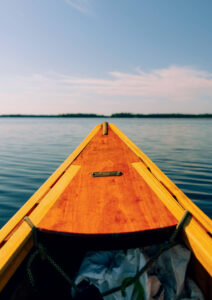 The front of a wooden canoe on calm water under a clear blue sky.