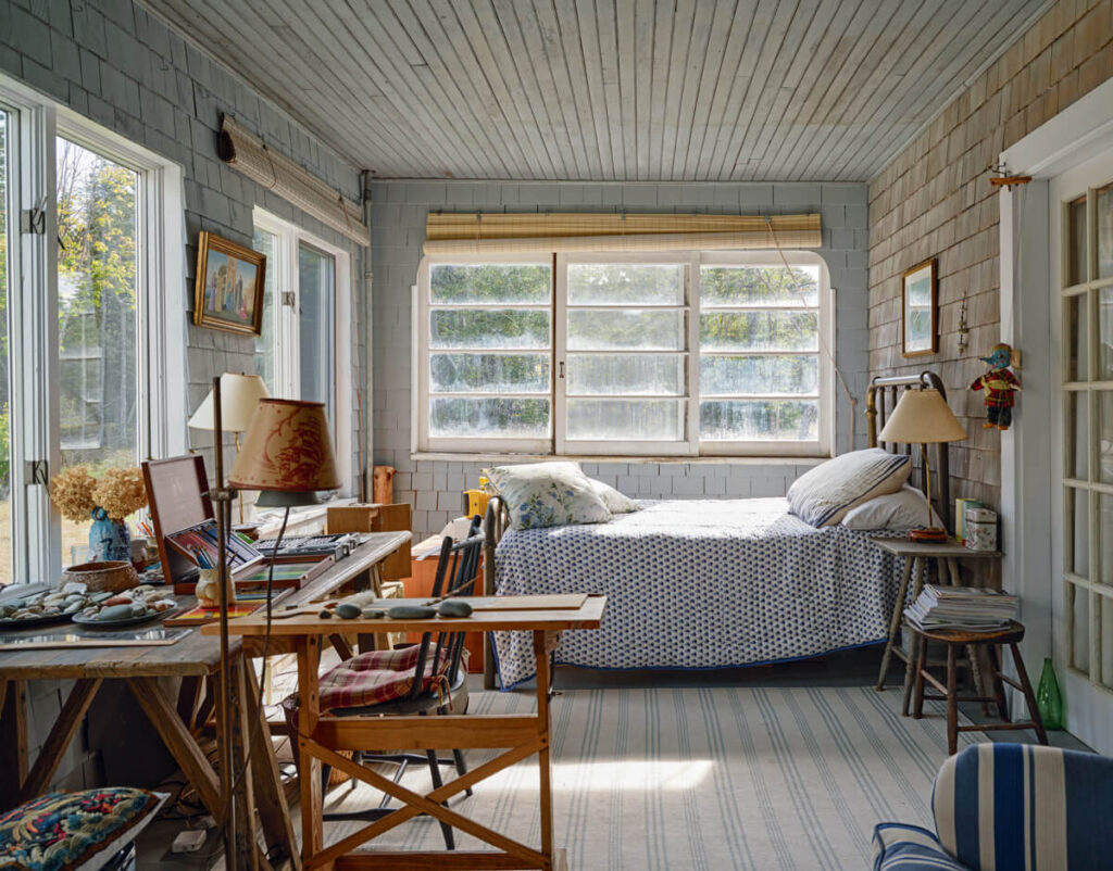 Sunlit cozy room with a bed by large windows, desk, chairs, and various decor and art supplies.