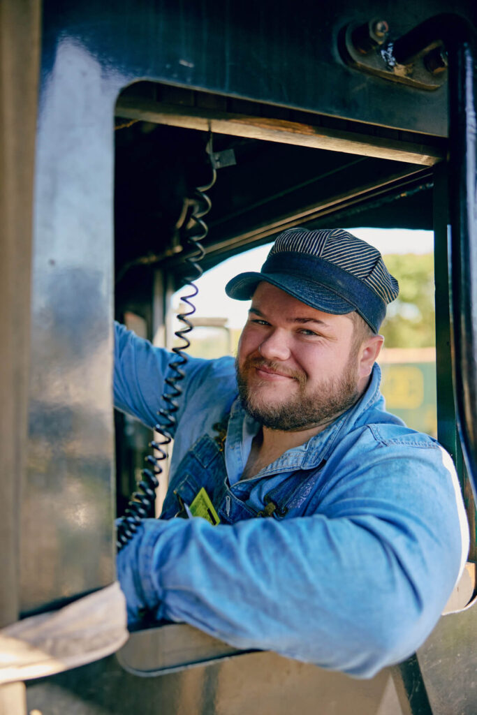 All Aboard the Essex Steam Train & Riverboat in Essex, Connecticut. Man in denim overalls and cap smiling while sitting in the cab of a locomotive, holding a radio handset.