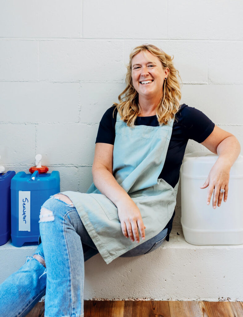 Smiling woman in apron sits between large blue and white containers against a white wall on a wooden floor.