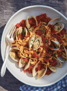 Tagliatelle pasta with clams in tomato sauce, garnished with herbs, served on a white plate with a fork and spoon.