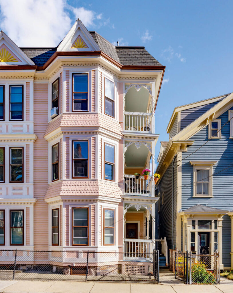 A pink Victorian-style house with bay windows next to a blue house under a clear sky.