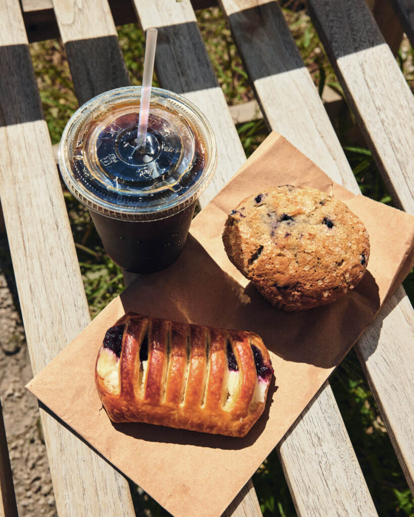 Iced coffee, a blueberry muffin, and a fruit-filled pastry on a brown paper, placed on a wooden bench.