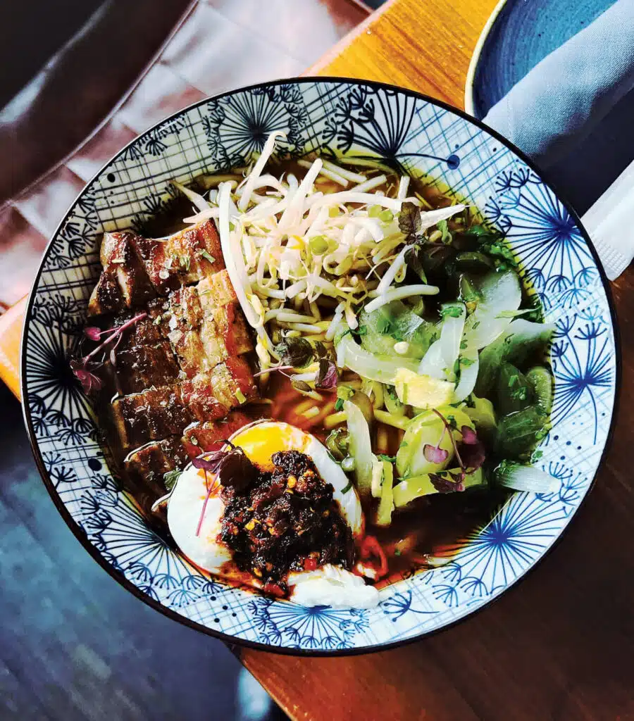 A bowl of ramen with sliced pork, soft-boiled egg, bean sprouts, and greens in a patterned bowl.