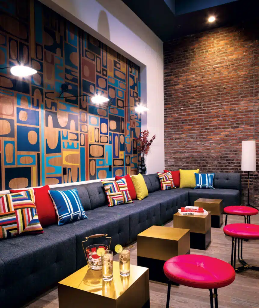 Modern lounge with a long gray sofa, colorful pillows, brick wall, geometric art, and gold tables with drinks.