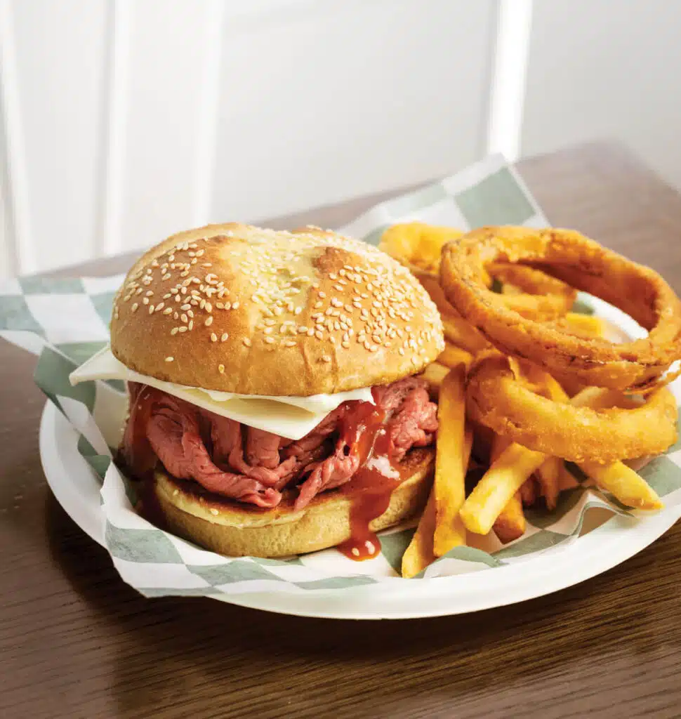 A roast beef sandwich with cheese, served with onion rings and fries on a paper-lined plate.