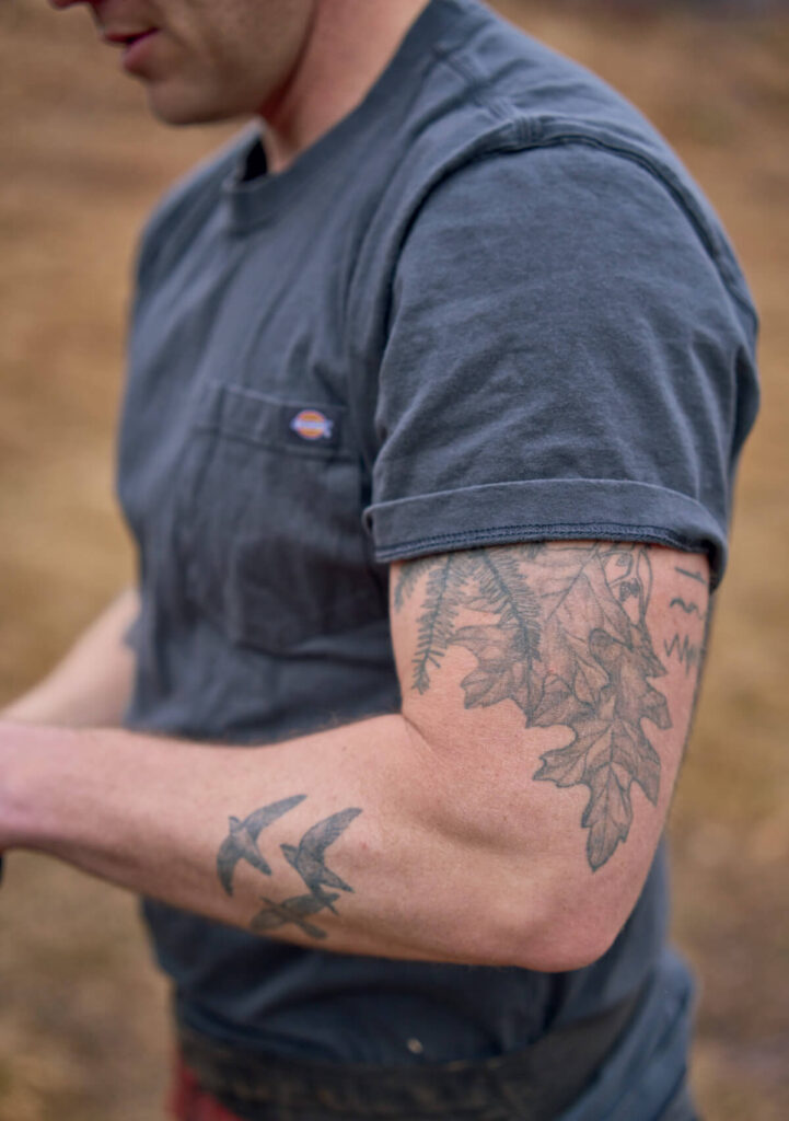 A man in a gray t-shirt shows arm tattoos of oak leaves, ferns, and birds.