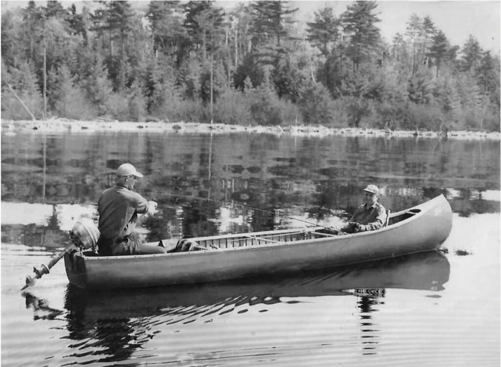 Two people in a canoe fishing on a calm lake, surrounded by trees and reflected in the water.