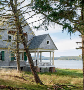 Gray shingled house with a covered porch, facing a lake, surrounded by trees and grassy lawn.