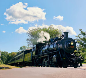 Black steam locomotive pulls vintage green passenger cars on a sunny day with blue sky and scattered clouds.