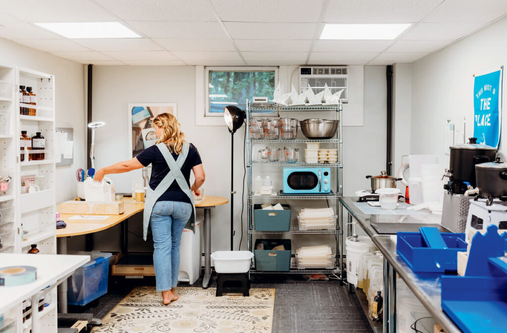 A woman in an apron works in a well-organized, brightly lit home workshop with various supplies and equipment.