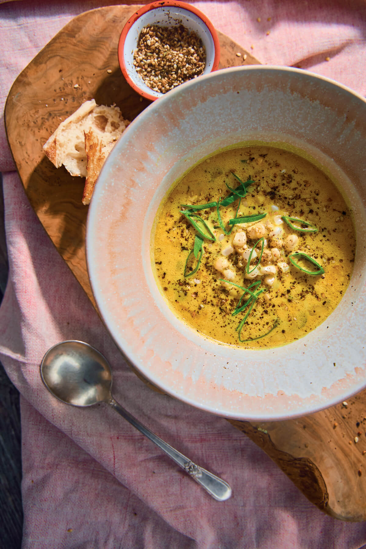 Sweet Corn Vegetarian Chowder. Bowl of yellow soup with herbs, bread, and seasoning on a wooden board with a spoon on a pink cloth.