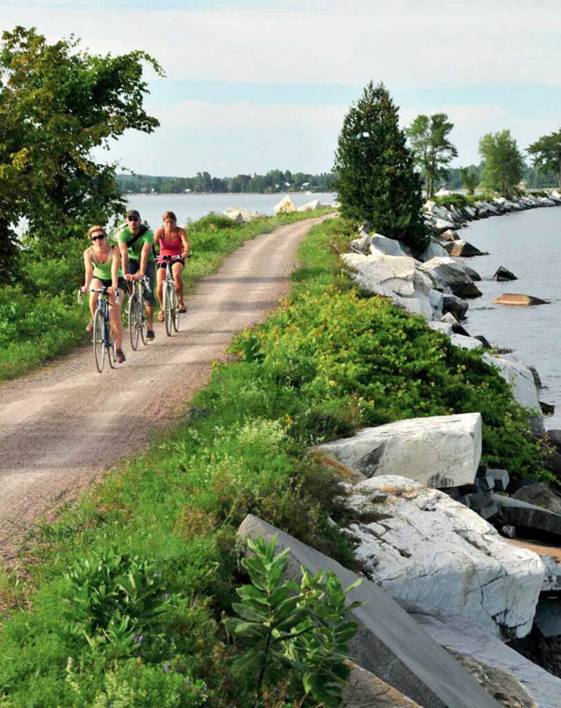 Three people ride bikes on a gravel path beside a rocky shoreline with greenery and trees.