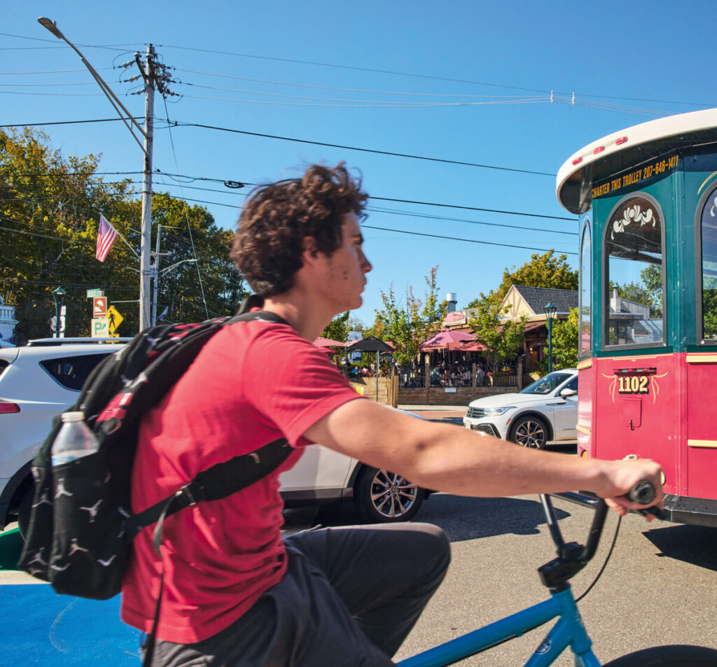 A man with a backpack rides a bicycle past a red trolley on a sunny street with cars and trees.