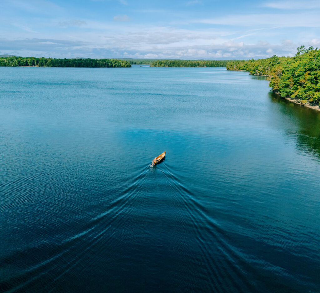 A canoe with two people rows across a calm blue lake surrounded by trees under a partly cloudy sky.