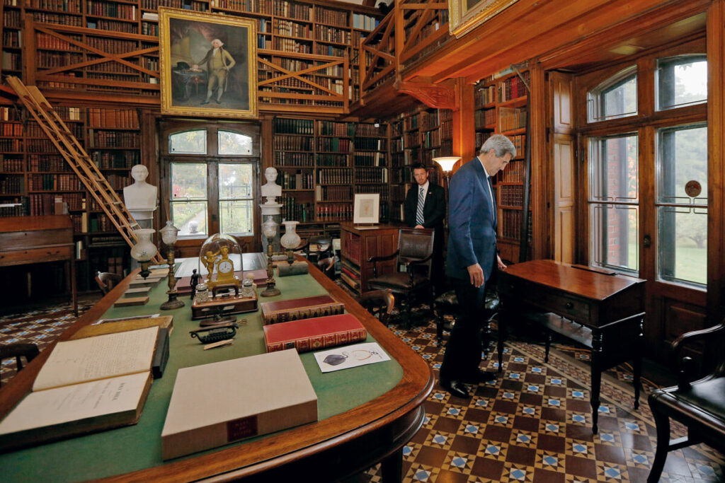 Two men in a historic library room with bookshelves, a large desk, busts, and checkered tile floor.
