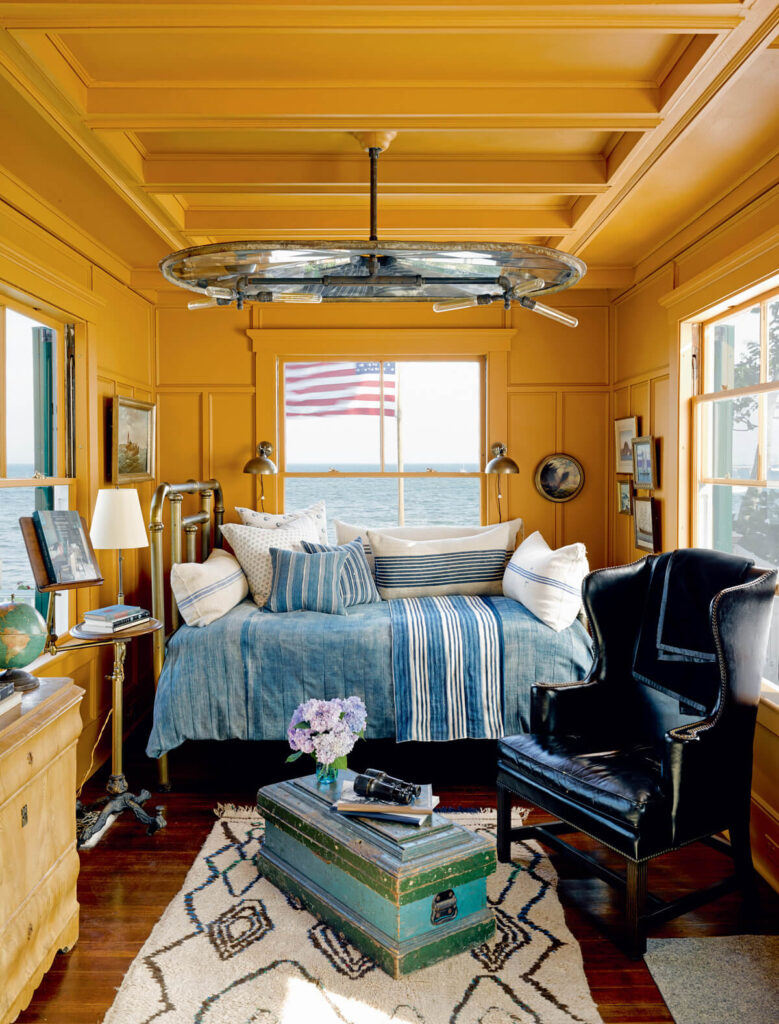 Sunny yellow-paneled room with a striped bed, black armchair, vintage trunk, and a window view of water and a flag.