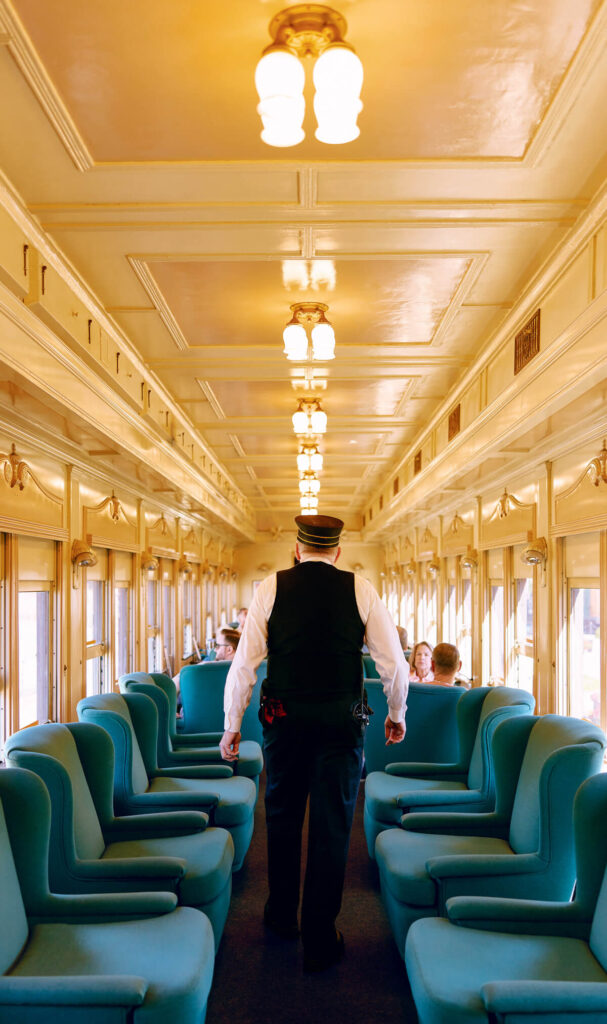 Train conductor walking down an aisle between teal seats in a vintage, well-lit train car.