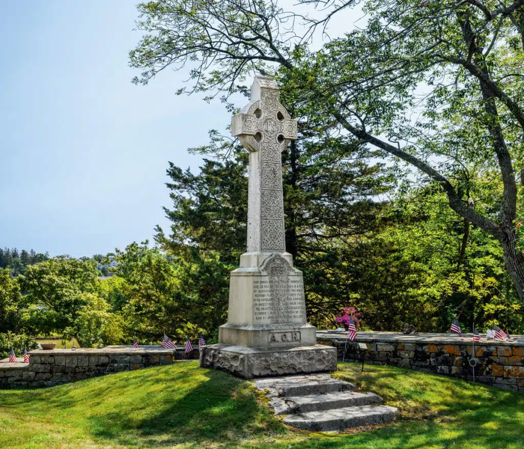 Celtic cross memorial with inscriptions, surrounded by American flags and greenery under a clear sky.