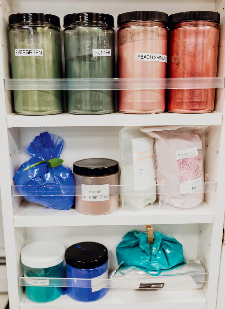 Jars and bags of colored powders and clays are organized on white shelves, each labeled with their color or type.