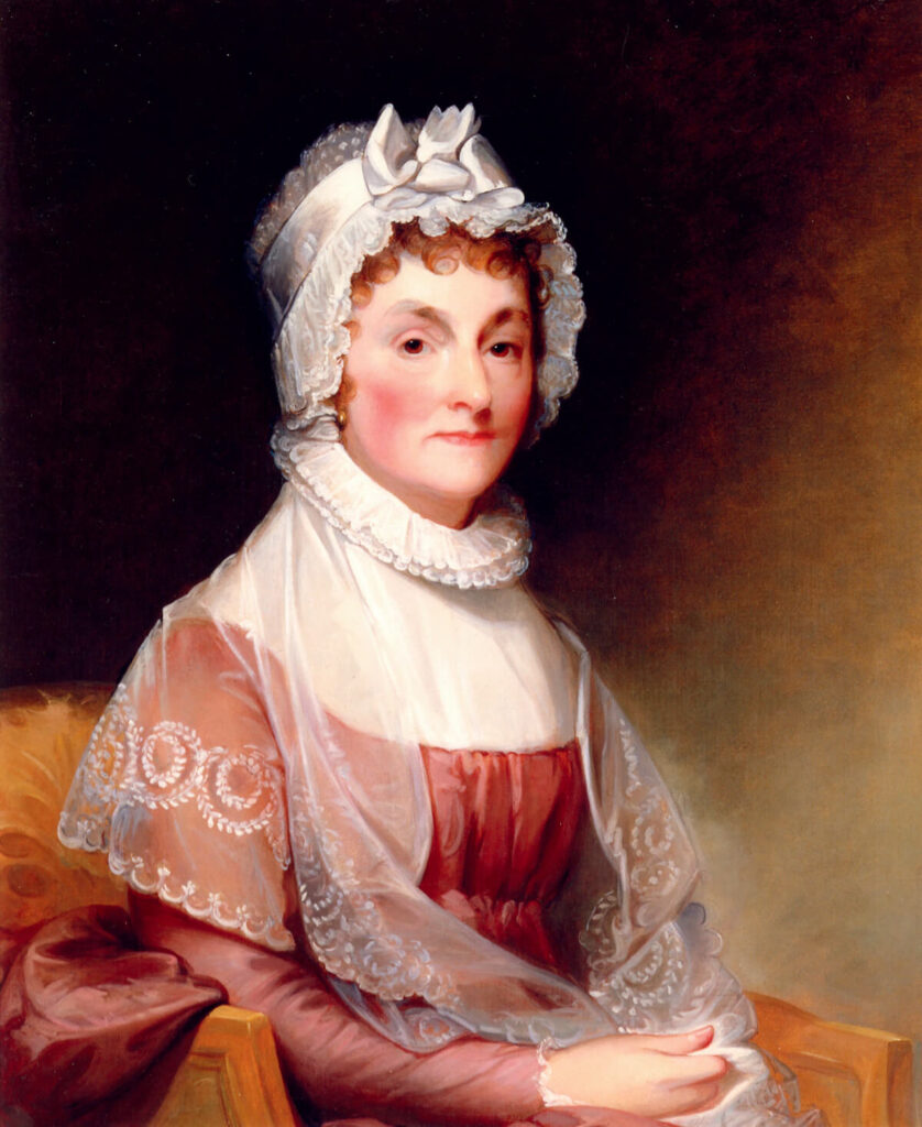 A woman in a lace bonnet and shawl sits on a chair, wearing a pink dress and looking forward calmly.